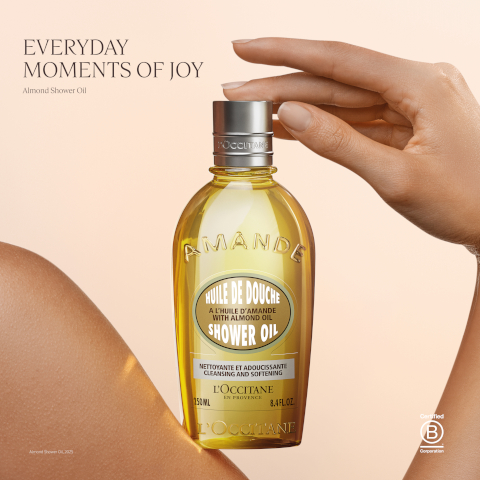 Image 1 - A bottle of LOccitane Almond Shower Oil, featuring a gold and clear design, is held by a hand with a light skin tone against a soft, neutral background; visible text includes EVERYDAY MOMENTS OF JOY, Amande, HUILE DE DOUCHE, SHOWER OIL, and L Occitane.